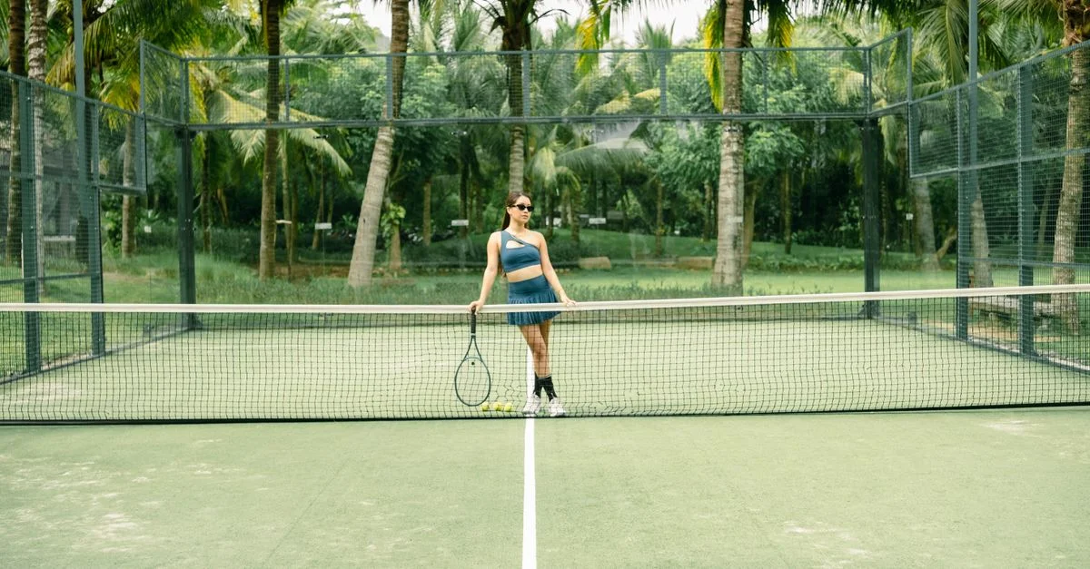 Mujer con atuendo de tenis de pie en una cancha verde, sosteniendo una raqueta.
