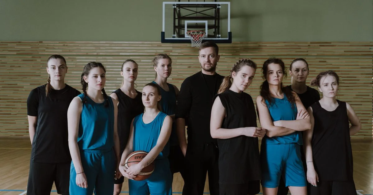 Un equipo femenino de baloncesto posando con su entrenador dentro de un gimnasio.