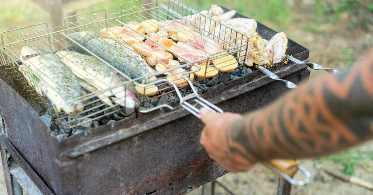 Pescado y carnes a la parrilla en una barbacoa con un brazo tatuado manejando la parrilla al aire libre.