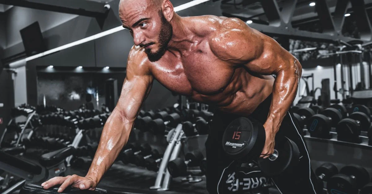 Un hombre en forma y musculoso haciendo ejercicio sin camiseta con mancuernas en un entorno de gimnasio, mostrando fuerza y concentración.
