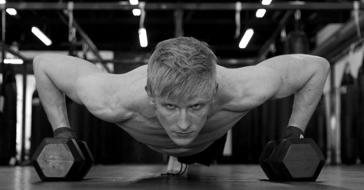 Imagen en blanco y negro de un hombre concentrado haciendo flexiones con pesas en un gimnasio.