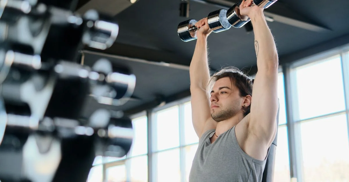 Hombre realizando press de hombros con mancuernas en un gimnasio bien iluminado.