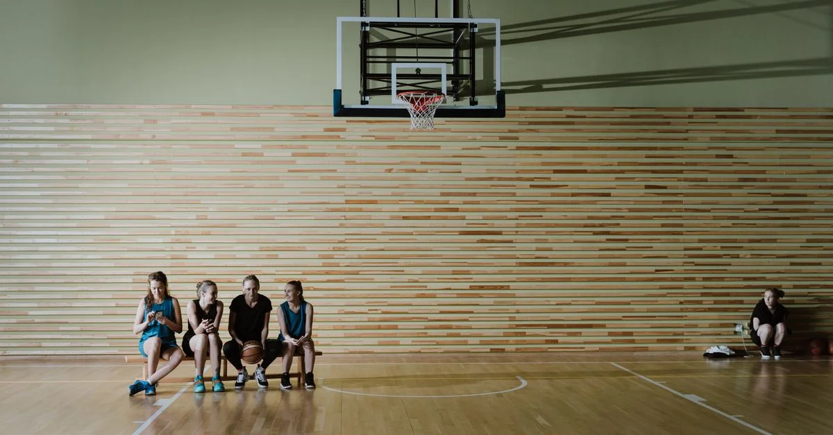Jugadoras jóvenes de baloncesto sentadas en un banco del gimnasio debajo de un aro de baloncesto, descansando después de la práctica.
