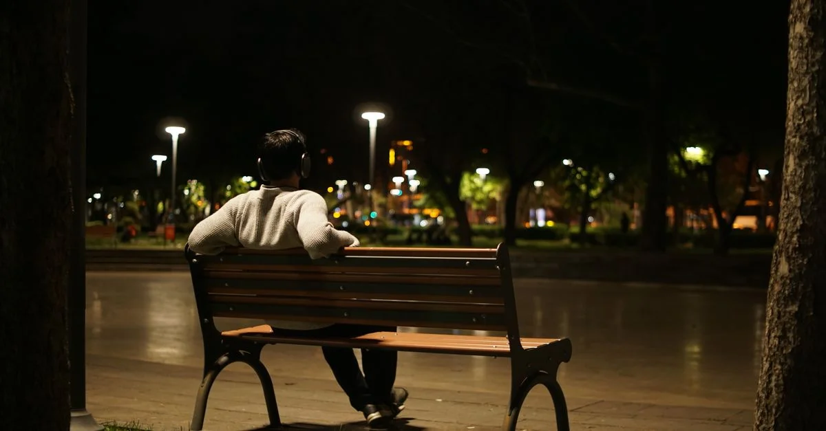 Un hombre con auriculares sentado solo en un banco del parque bajo las farolas por la noche.