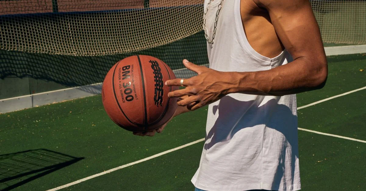 Primer plano de un hombre sosteniendo un balón de baloncesto en una cancha al aire libre, vistiendo una camiseta sin mangas.