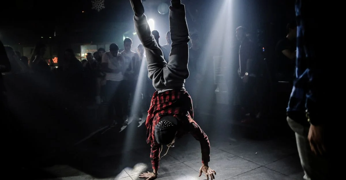 Un breakdancer en chemise à carreaux effectue un poirier dans une ambiance de club animée.