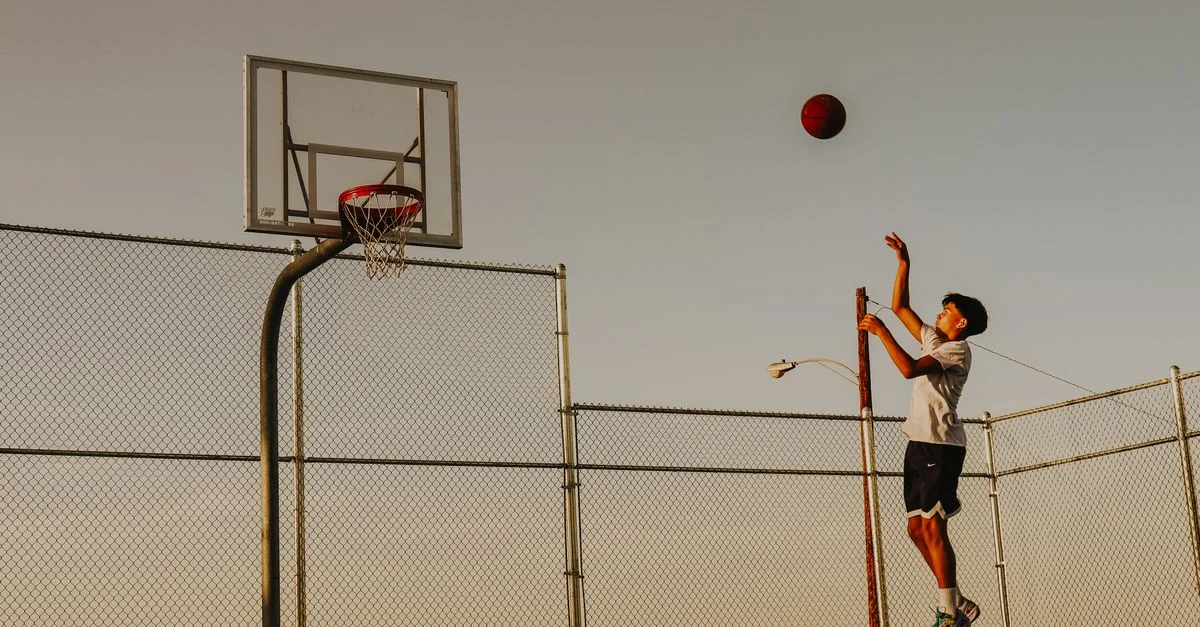 Niño adolescente jugando baloncesto al aire libre, tirando al aro con la puesta de sol.