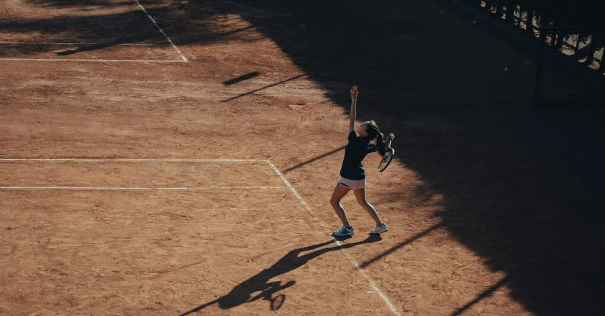 Vista aérea de una mujer jugando al tenis, sacando en una pista de tierra batida bajo la luz del sol.