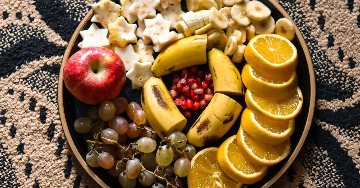 Plato colorido de frutas con plátanos, naranjas, manzana, uvas y granada sobre fondo decorativo.