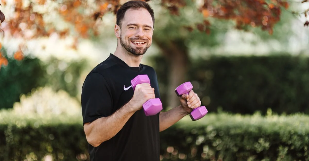 Hombre sonriente con camiseta negra haciendo ejercicio al aire libre con mancuernas moradas en un parque.