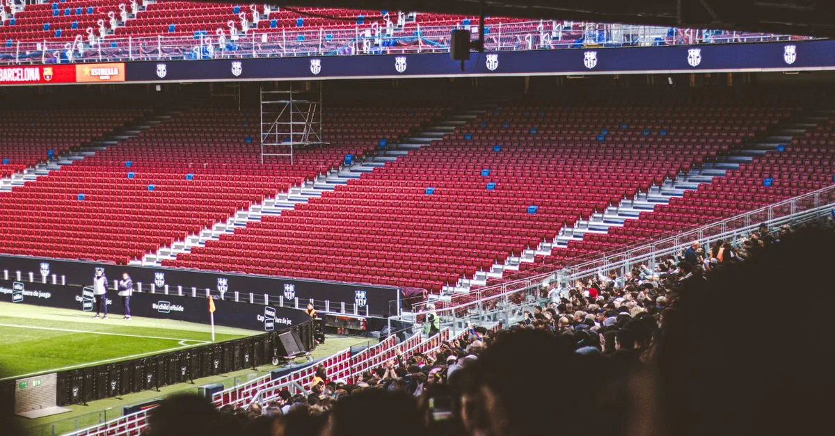 Vista panorámica del estadio con asientos rojos vacíos y público reunido para un partido en directo.