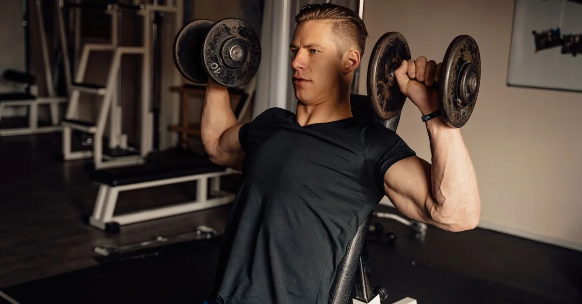 Un hombre concentrado en un gimnasio realizando un press de hombros con mancuernas, mostrando fitness y fuerza.