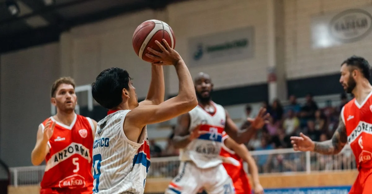 Partido dinámico de baloncesto en interiores con jugadores en plena acción, destacando trabajo en equipo y competencia.