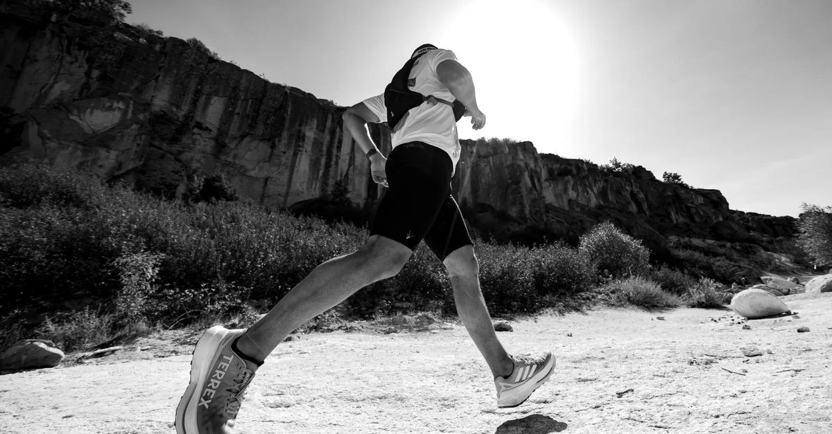 Fotografía en blanco y negro de un corredor de senderos debajo de un acantilado soleado, que muestra movimiento y determinación.