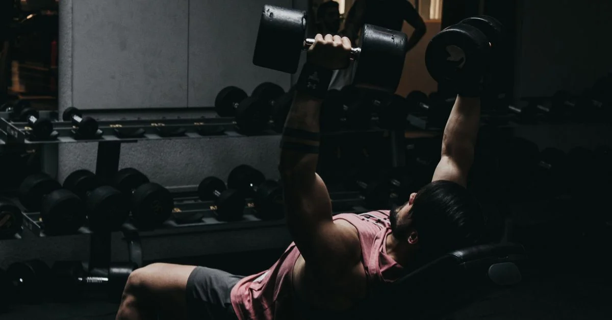 Hombre musculoso con ropa deportiva realizando press de banca con mancuernas en un gimnasio poco iluminado.