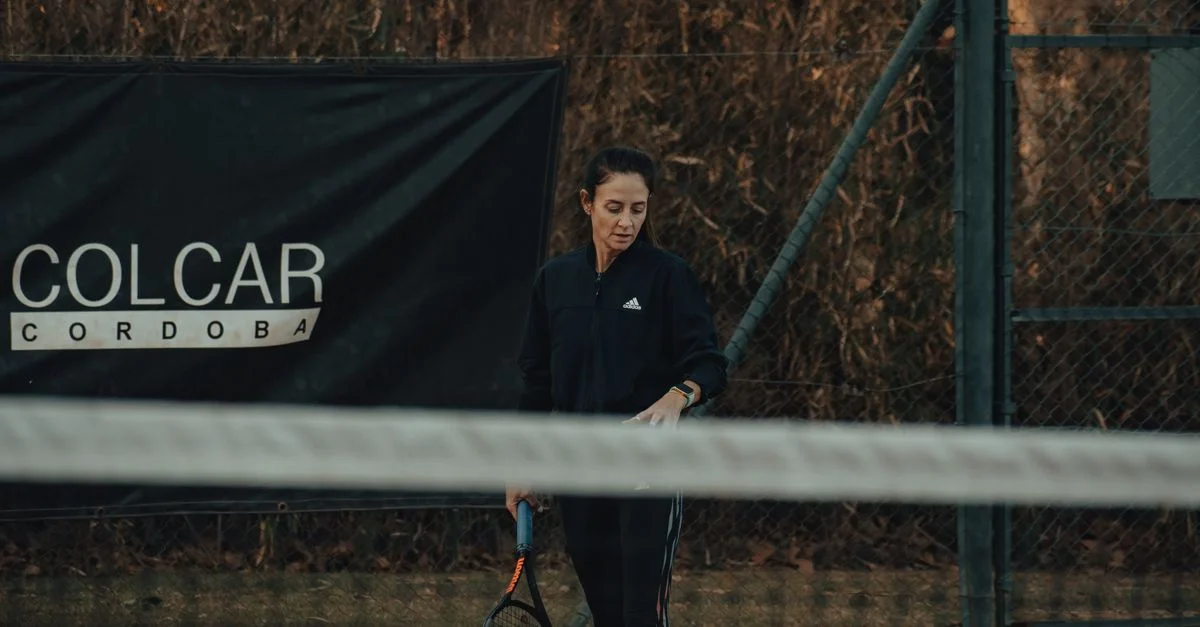 Una mujer sosteniendo una raqueta de tenis en una cancha de tenis de tierra batida al aire libre, junto a un cartel de COLCAR CORDOBA.